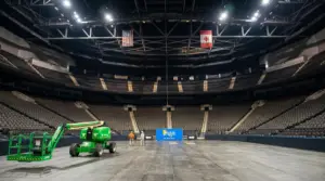 A wide view inside a large sports arena in Jacksonville, where two workers from General Sign Service Corporation use a green boom lift to safely install a massive blue Daily's sign.