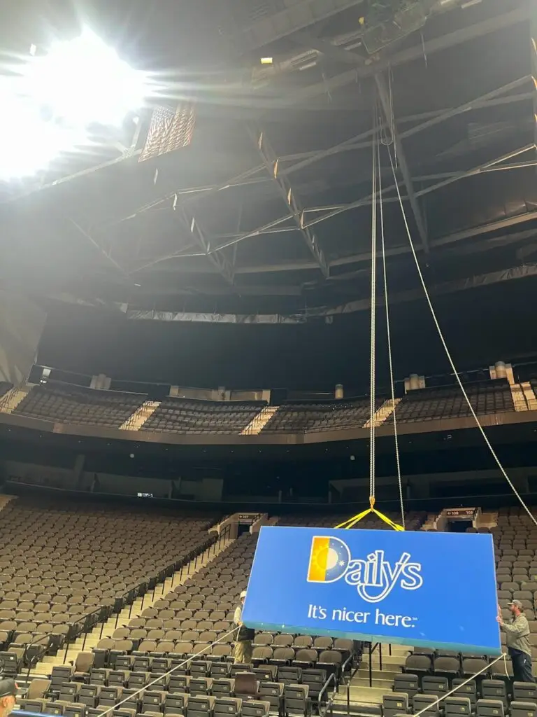 High-Stakes Branding: Engineering the 120-Foot Signage Installation at VyStar Memorial Arena 7 A large blue "Daily's" sign with the slogan "It's nicer here." being hoisted by chains and guide ropes inside a large, empty arena with tiered seating.